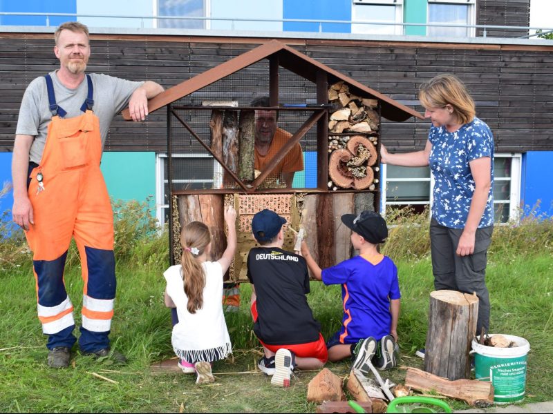 Zwei Erwachsene und drei Kinder befüllen ein großes Insektenhotel mit Naturmaterialien.