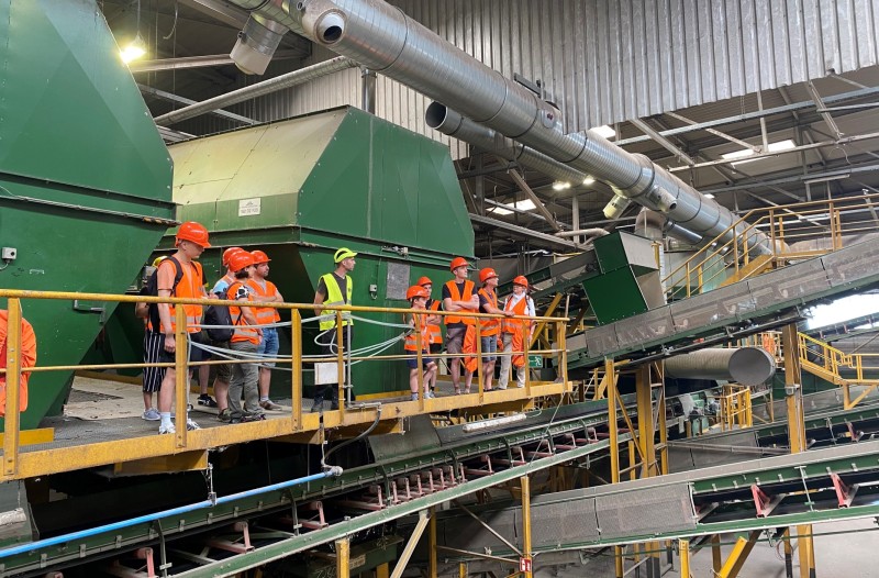 Personengruppe mit Arbeitsschutzbekleidung streht in einer Halle mit Förderbändern und verscheidenen Trennaggregaten.